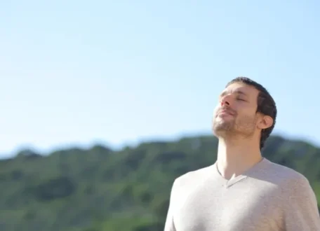 Man with eyes closed and face tilted toward the sun, breathing calmly outdoors with green hills behind him.