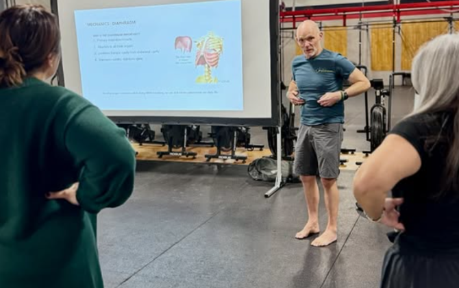 Barefoot instructor in a gym explains diaphragm mechanics to a small group, with an anatomy slide on a screen behind him.