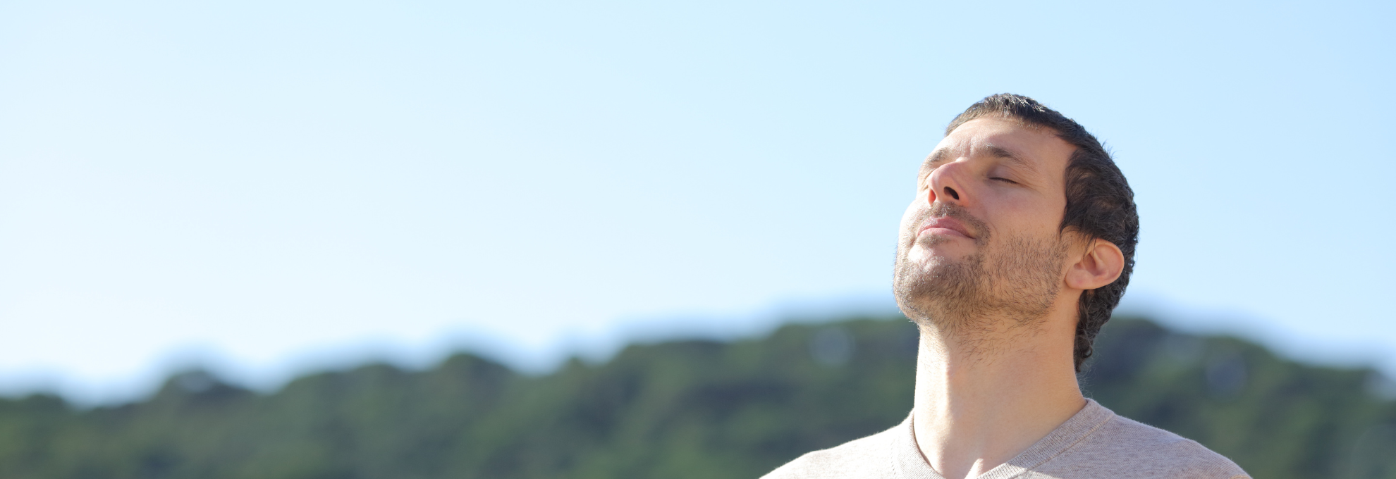 Man with eyes closed and face tilted toward the sun, breathing calmly outdoors with green hills behind him.
